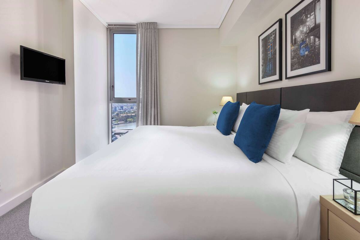 Ein Hotelzimmer mit einem großen Bett, einem flachbildfernseher an der Wand, einem Fenster mit Stadtblick und zwei gerahmten Bildern.