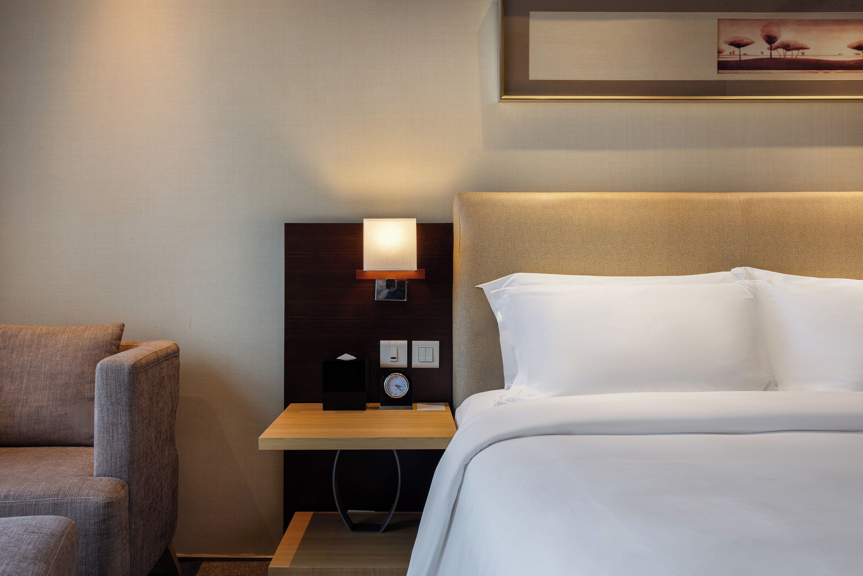 Een hotelkamer met een bed, nachtkastje met lamp, klok en stoel.