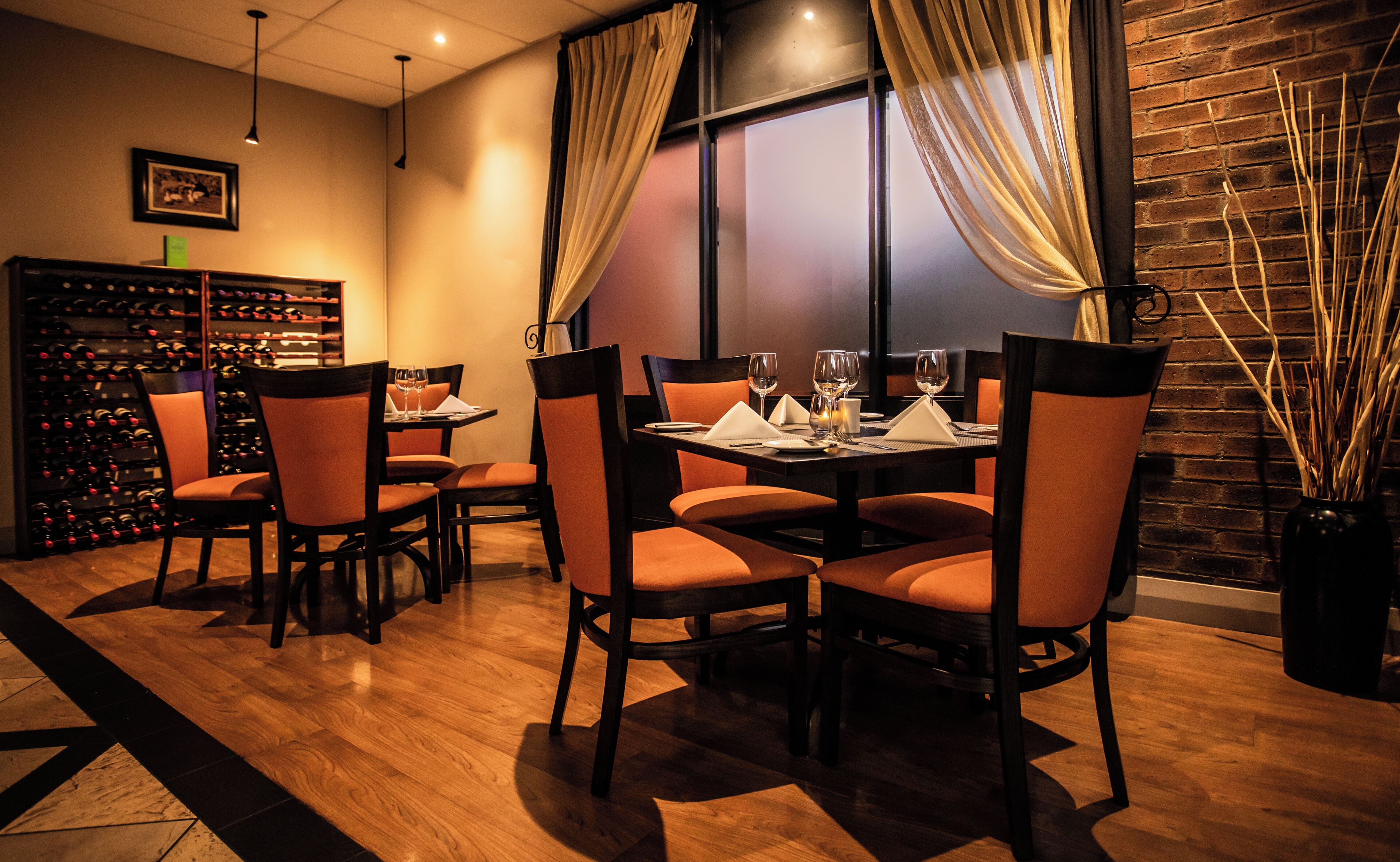 A dining area with a brick wall, wooden flooring, and orange chairs. There is a wine rack in the background and a window with curtains.