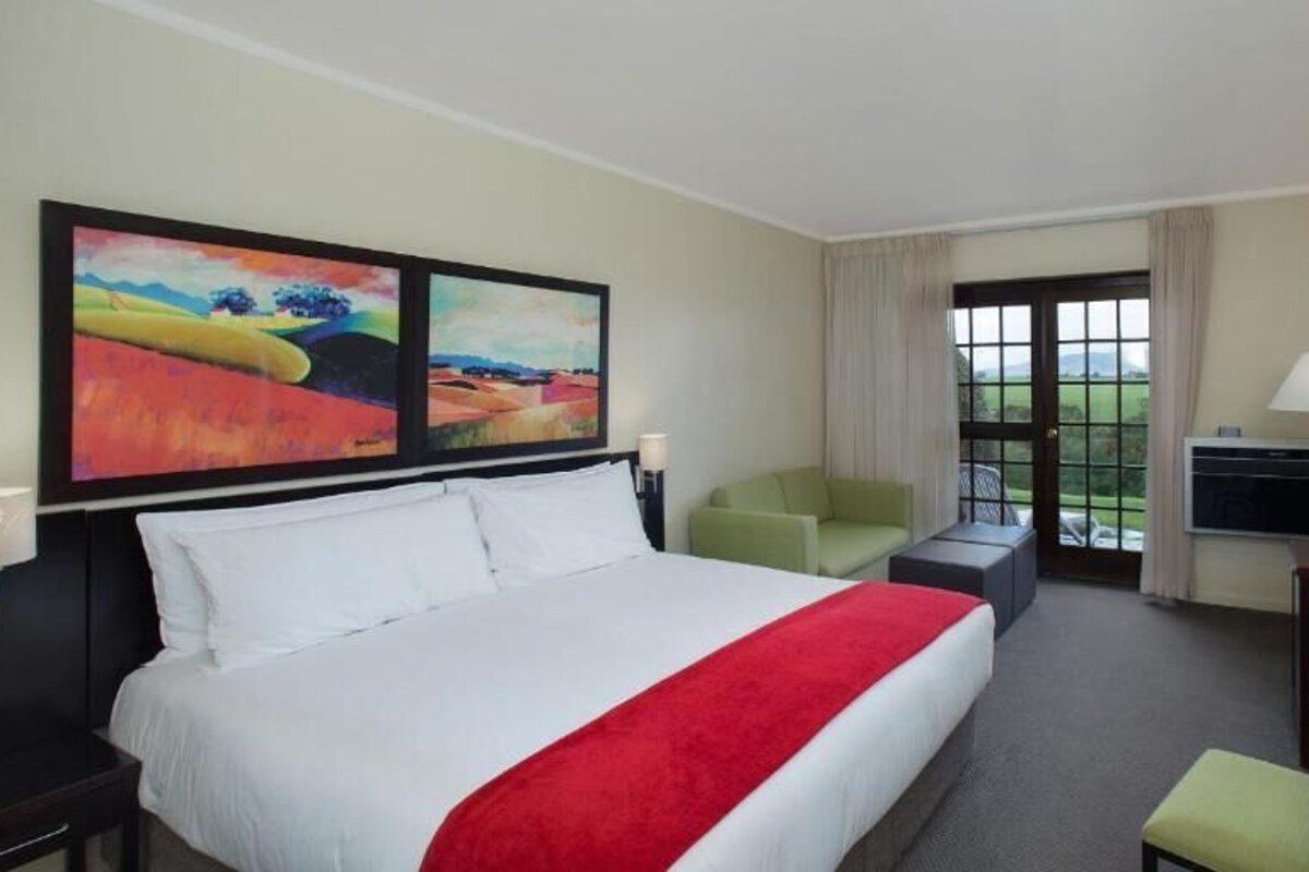 Une chambre d’hôtel avec un grand lit, un bureau, une chaise, deux tableaux au mur et une vue sur l’extérieur.
