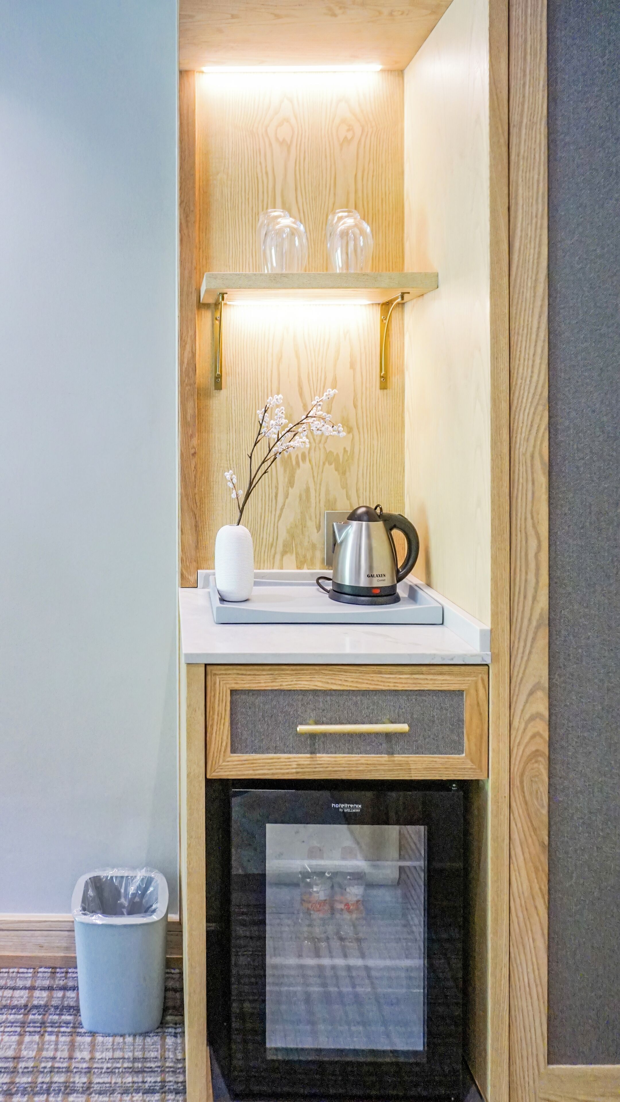 A compact kitchenette with a stainless steel kettle, a small vase with flowers, and a trash bin.