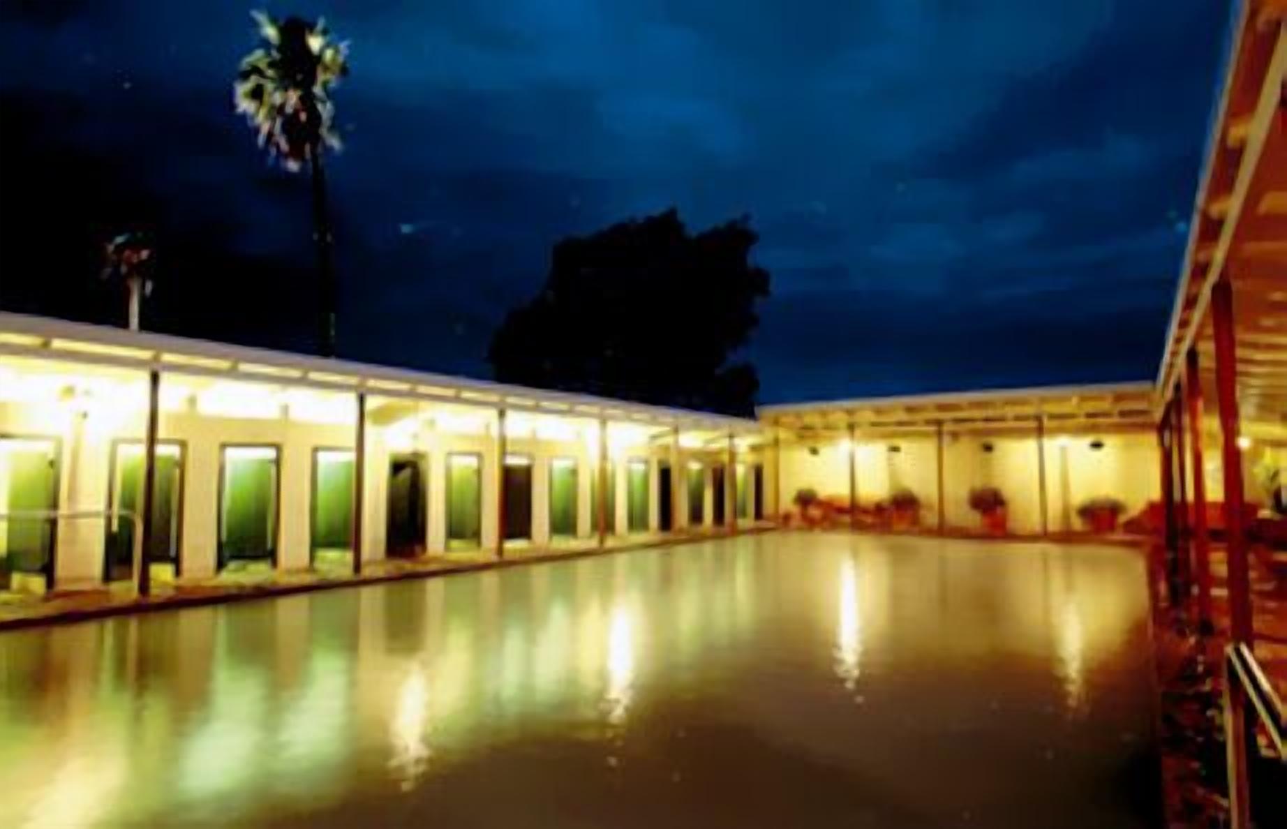 A nighttime view of a hotel with a swimming pool, reflecting lights, and a palm tree.