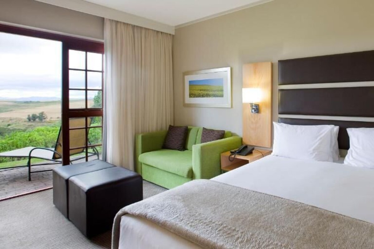 Une chambre d’hôtel avec un grand lit, un fauteuil vert, une petite table et une vue sur la campagne.