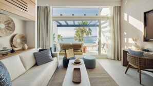 A modern living room with a sofa, ottoman, and coffee table, a wicker chair, and a view of the beach through large windows.