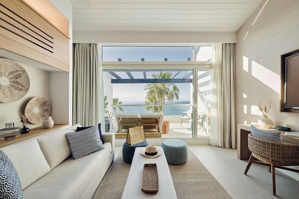 Ein modernes Wohnzimmer mit einer Couch, Hocker und einem Couchtisch, einem Korbstuhl und Blick auf den Strand durch große Fenster.