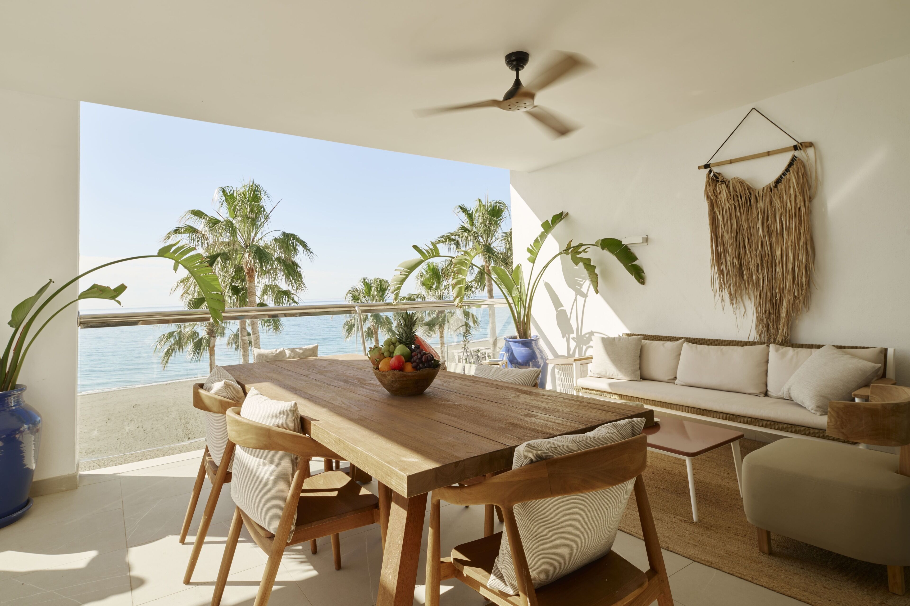 A wooden dining table with chairs, a sofa, and a ceiling fan in a room with a view of the sea.