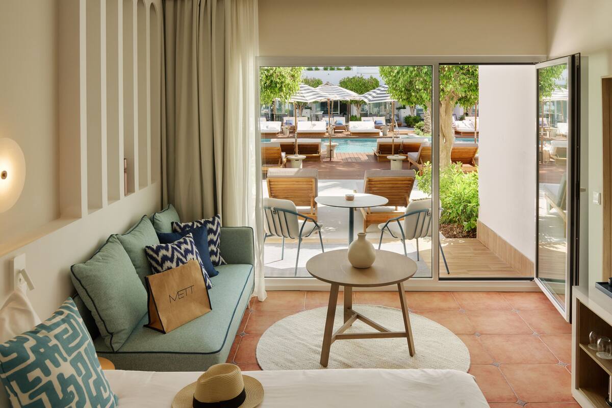 A modern hotel room with a sofa, a dining table, and a view of an outdoor pool area.