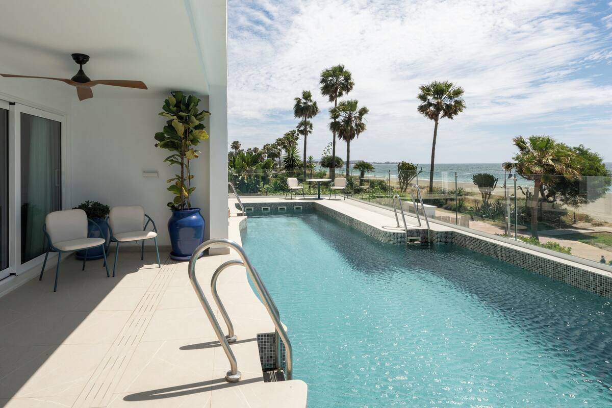 A swimming pool with a view of the beach, palm trees, and a ceiling fan.