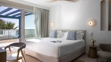 A modern hotel room with a large bed, a ceiling fan, a view of the ocean, and a balcony with a pool.