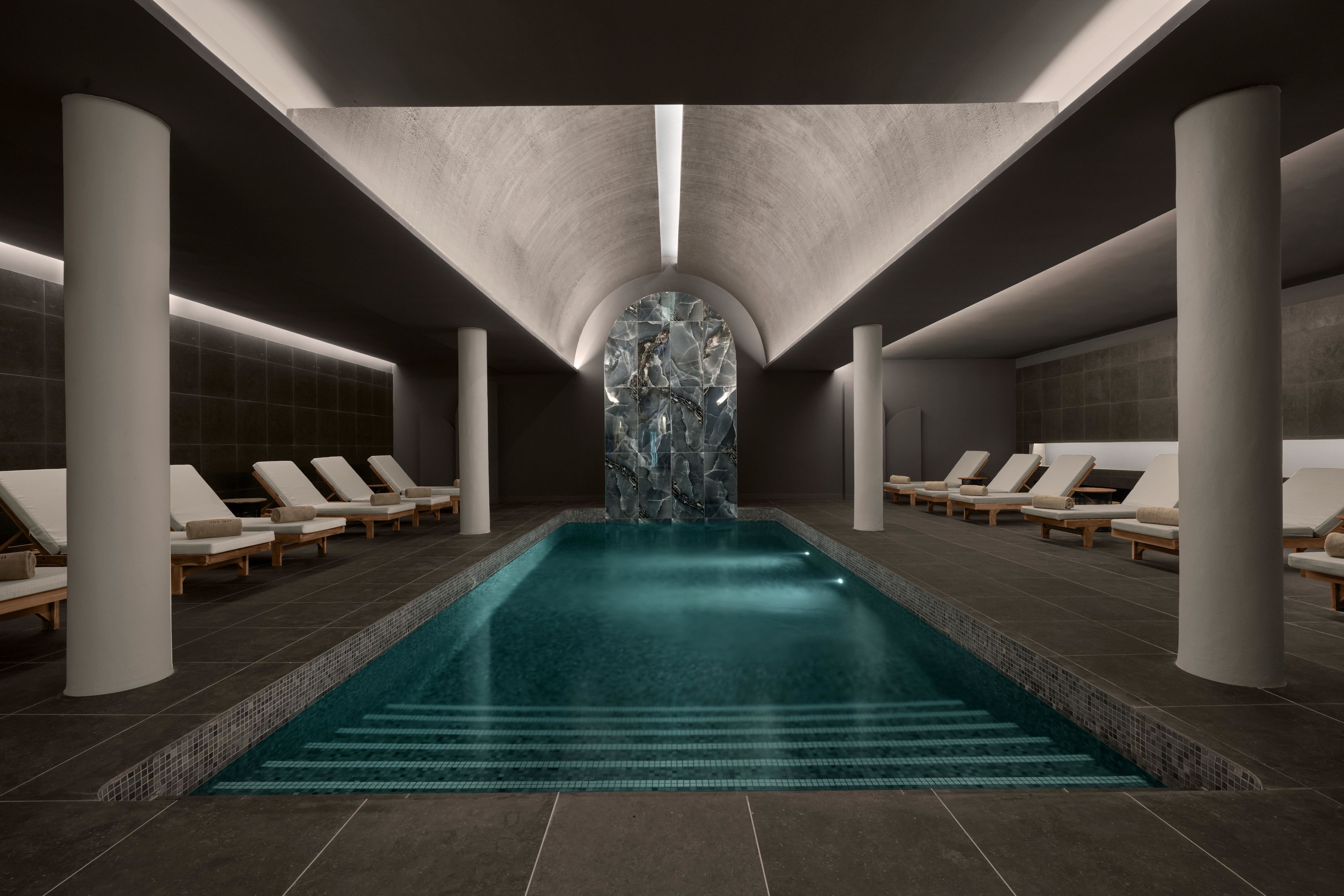 A modern indoor swimming pool with a central water feature, surrounded by lounge chairs and a tiled floor.