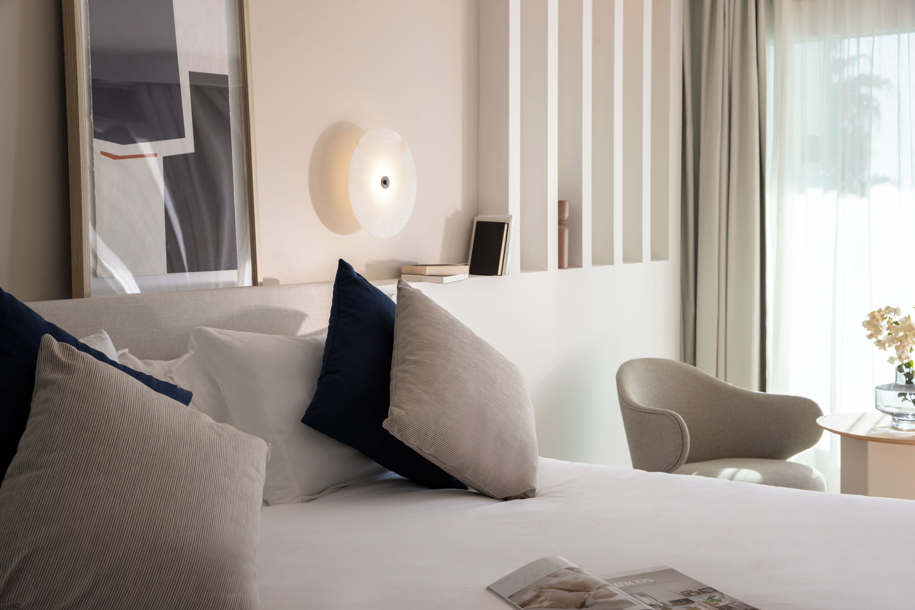 A hotel room with a neatly made bed, a side table with a vase, and a chair.
