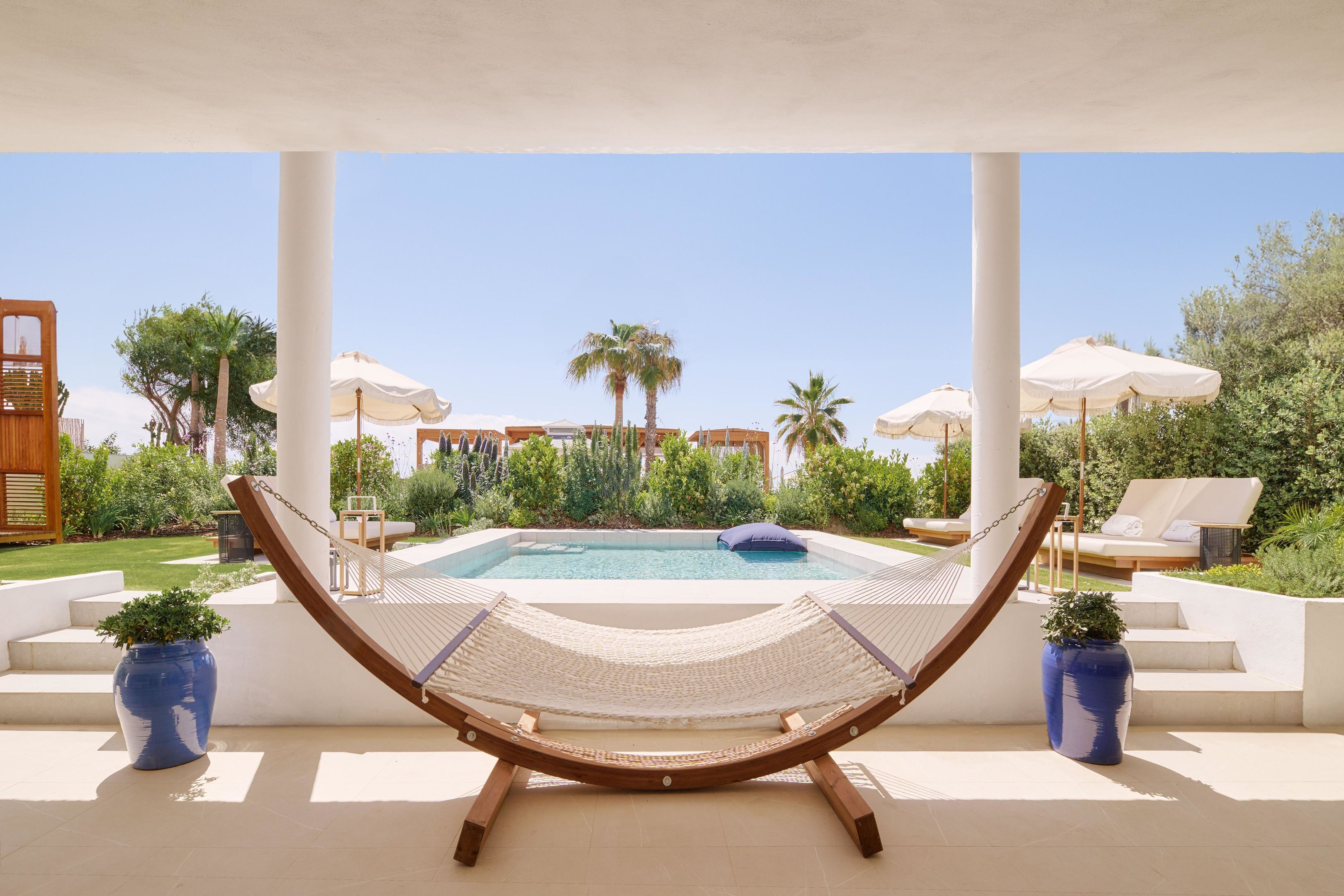 Una hamaca bajo un dosel blanco con vista a una piscina, rodeada de palmeras y sillas de descanso.