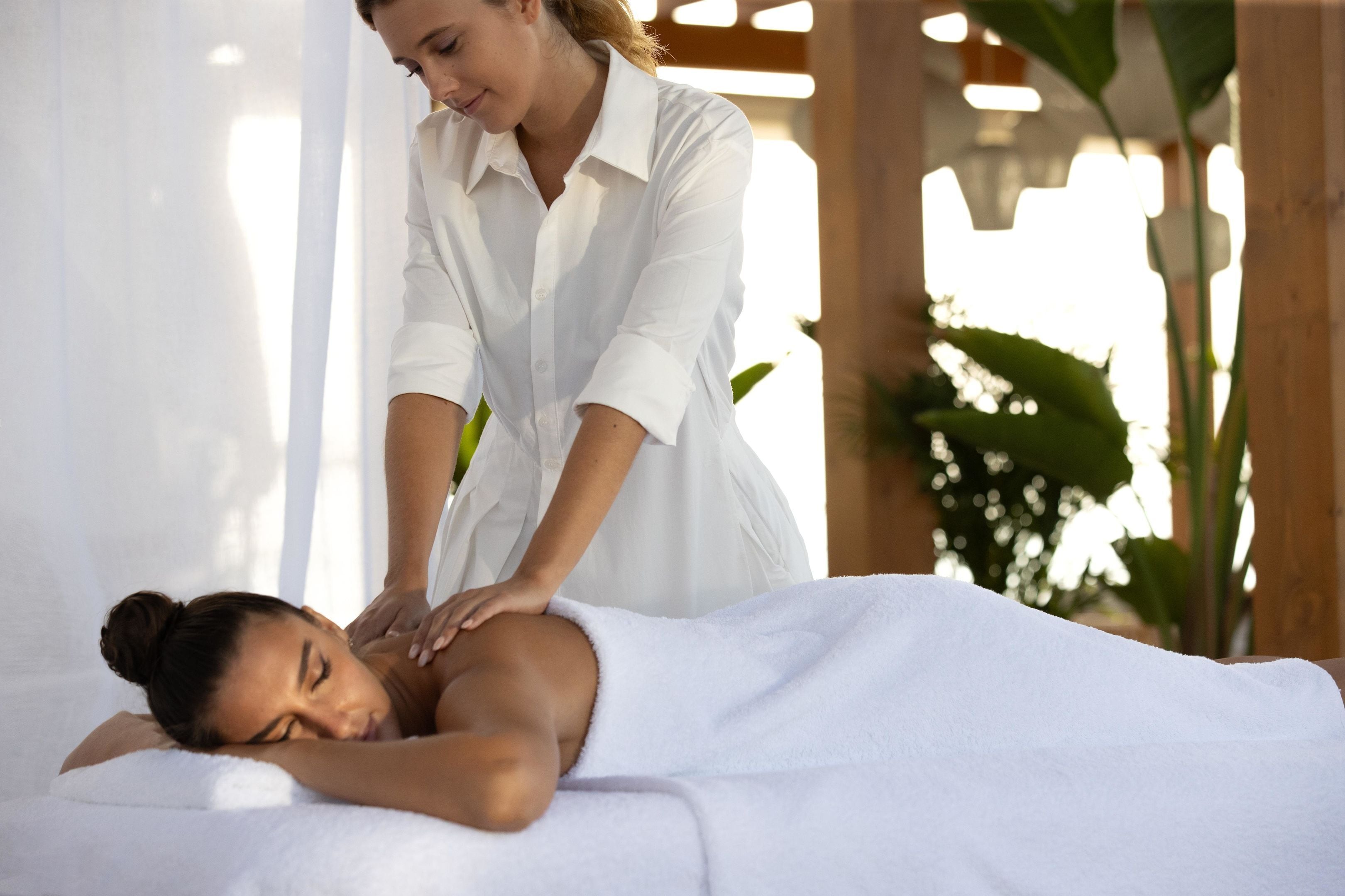 Sesión de masoterapia en un entorno de spa, con un cliente recostado en la camilla y un terapeuta atendiendo, además de una planta al fondo.
