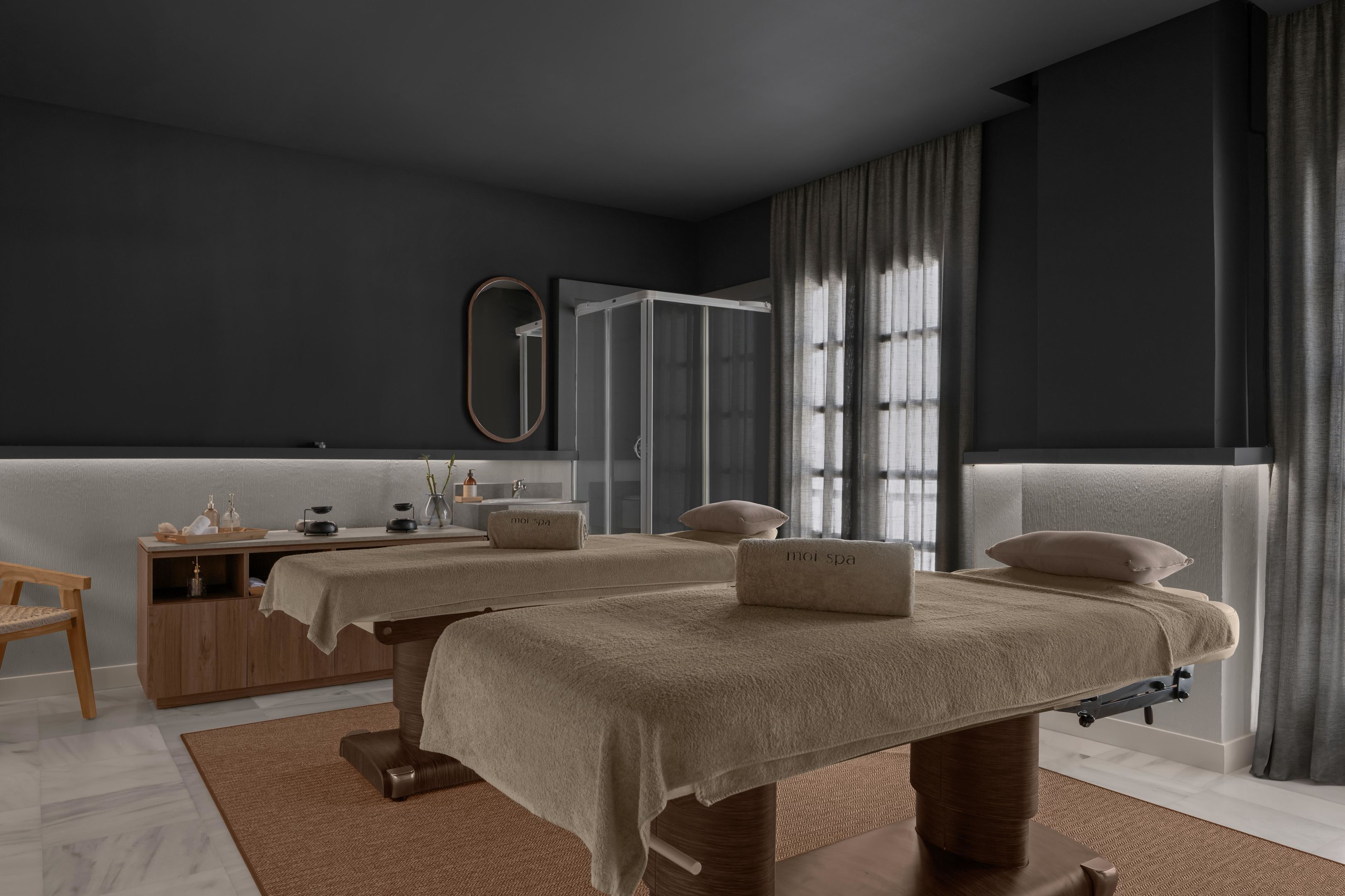 A modern spa room with two massage tables, a wooden vanity, and a glass shower enclosure.