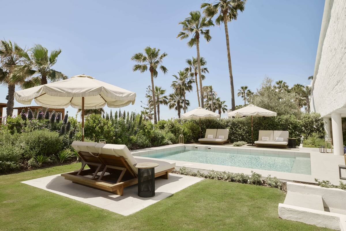 A pool area with lounge chairs and umbrellas, surrounded by palm trees and greenery.