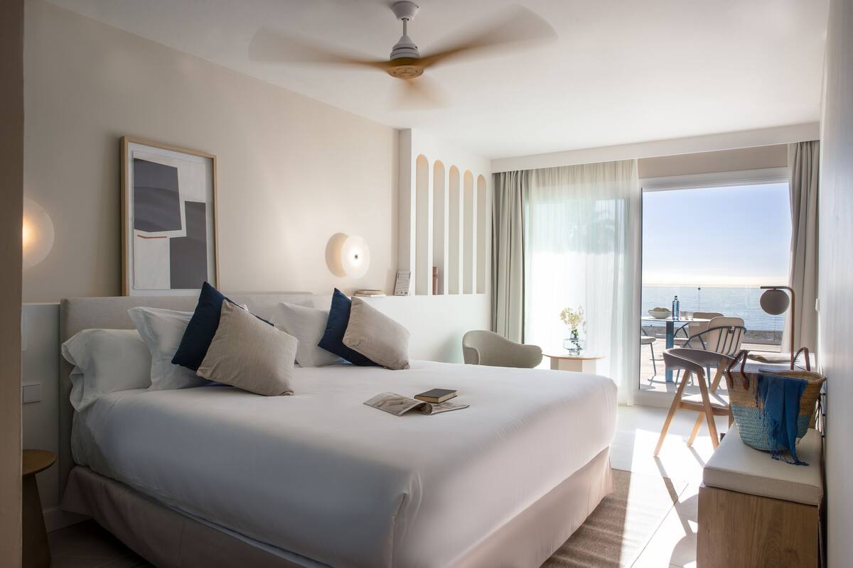 A hotel room with a large bed, a ceiling fan, a desk with a chair, and a view of the ocean.