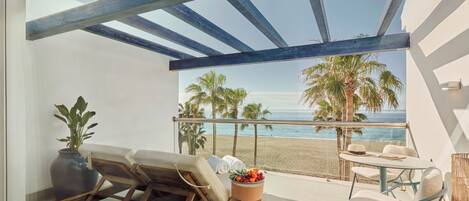 A balcony with a view of the beach, a seating area with cushions, and a table with a fruit bowl.