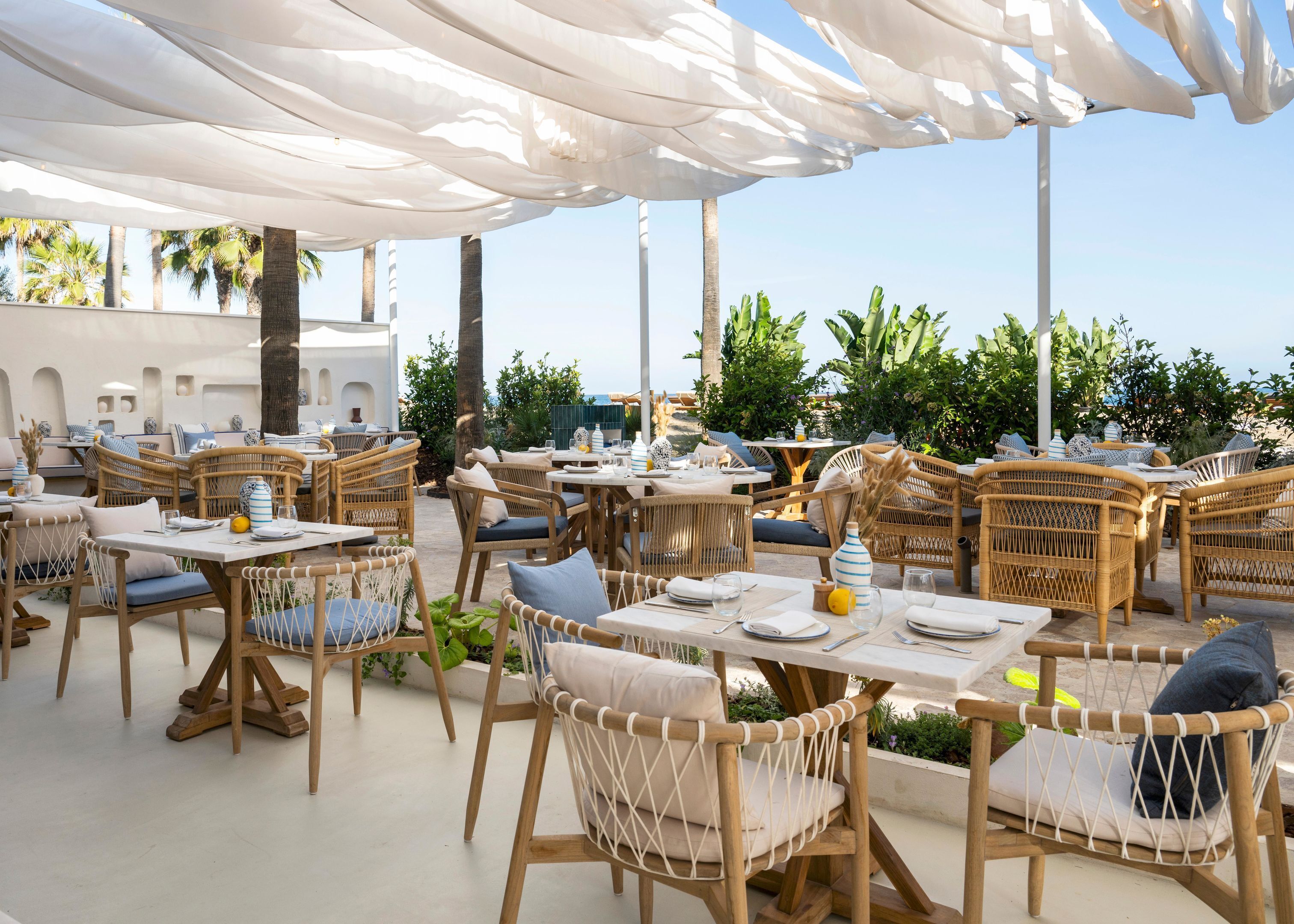 An outdoor dining area with wicker furniture and white canopies. Tables are set with plates, glasses, and cutlery.