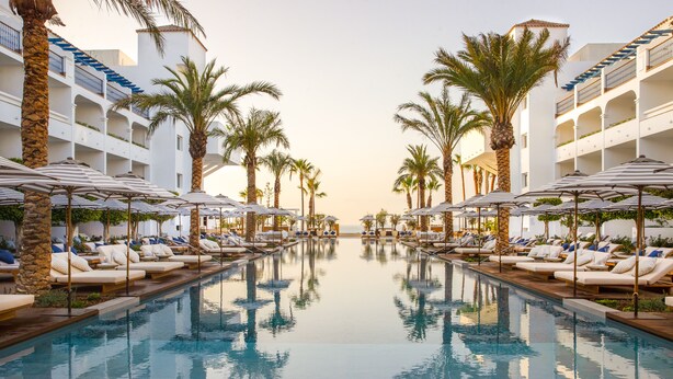 A pool area with lounge chairs and umbrellas, surrounded by palm trees and a building with balconies.