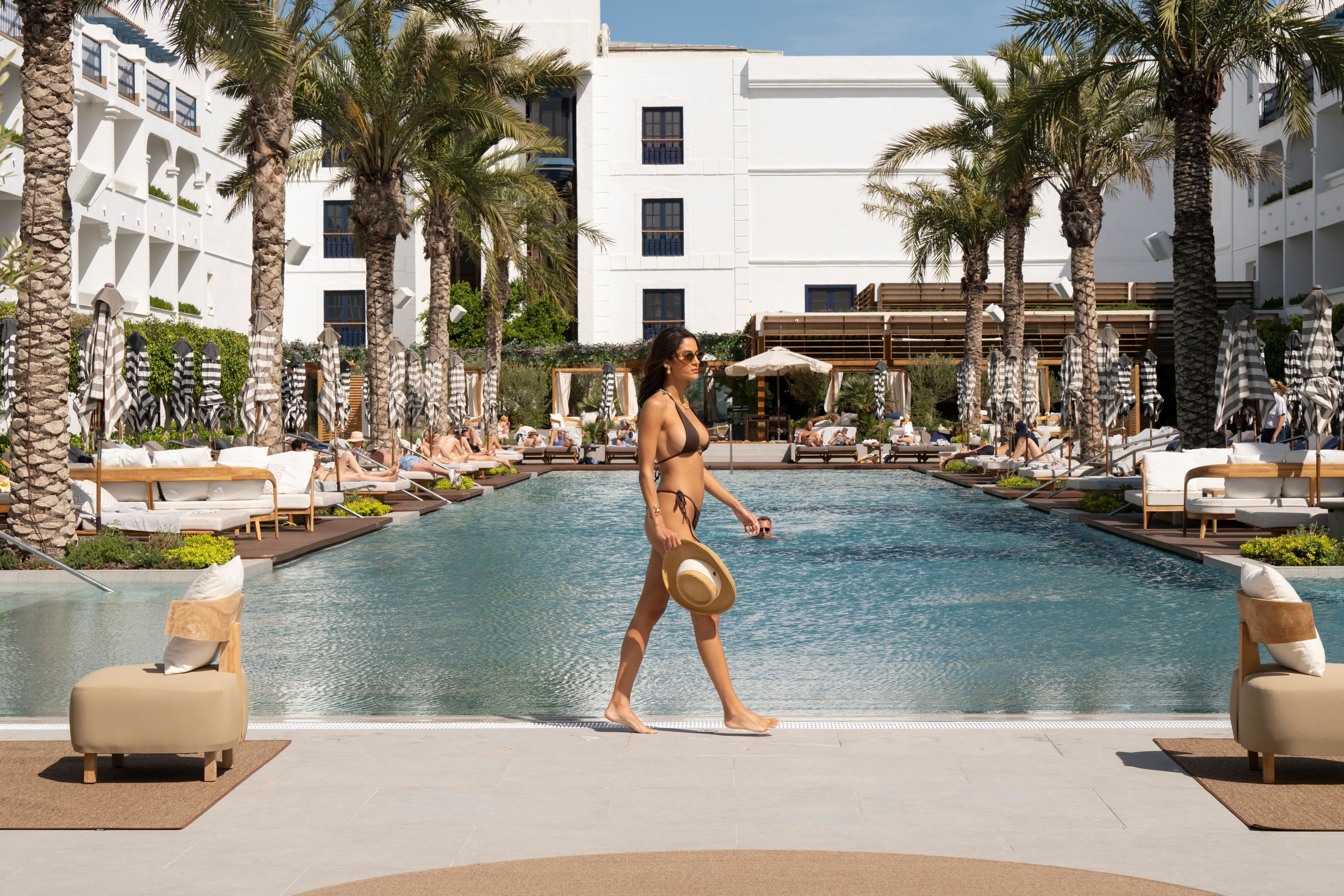 Un área de piscina con sillones de descanso y sombrillas, rodeada de palmeras y un edificio moderno.