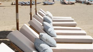 A row of sun loungers with patterned pillows and beige umbrellas on a sandy beach.