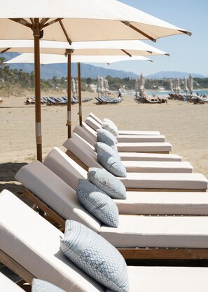 A row of sun loungers with patterned pillows and beige umbrellas on a sandy beach.