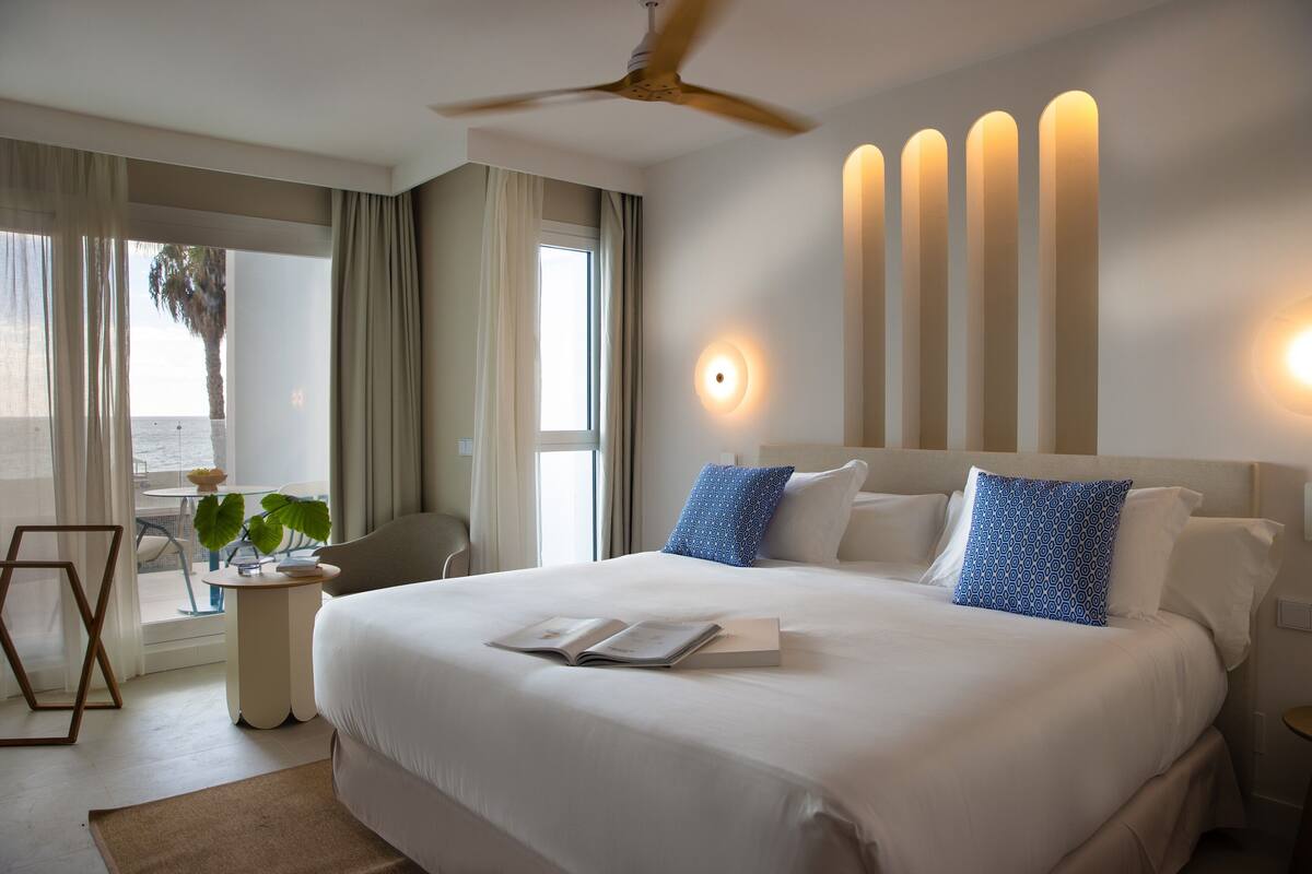 A hotel room with a large bed, a ceiling fan, a bedside table with a book, and a view of the ocean through the window.