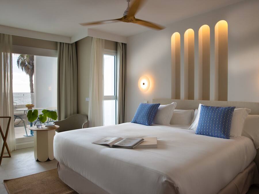 Habitación de hotel con una cama grande, un ventilador de techo, una mesita de noche con un libro y vista al océano por la ventana.