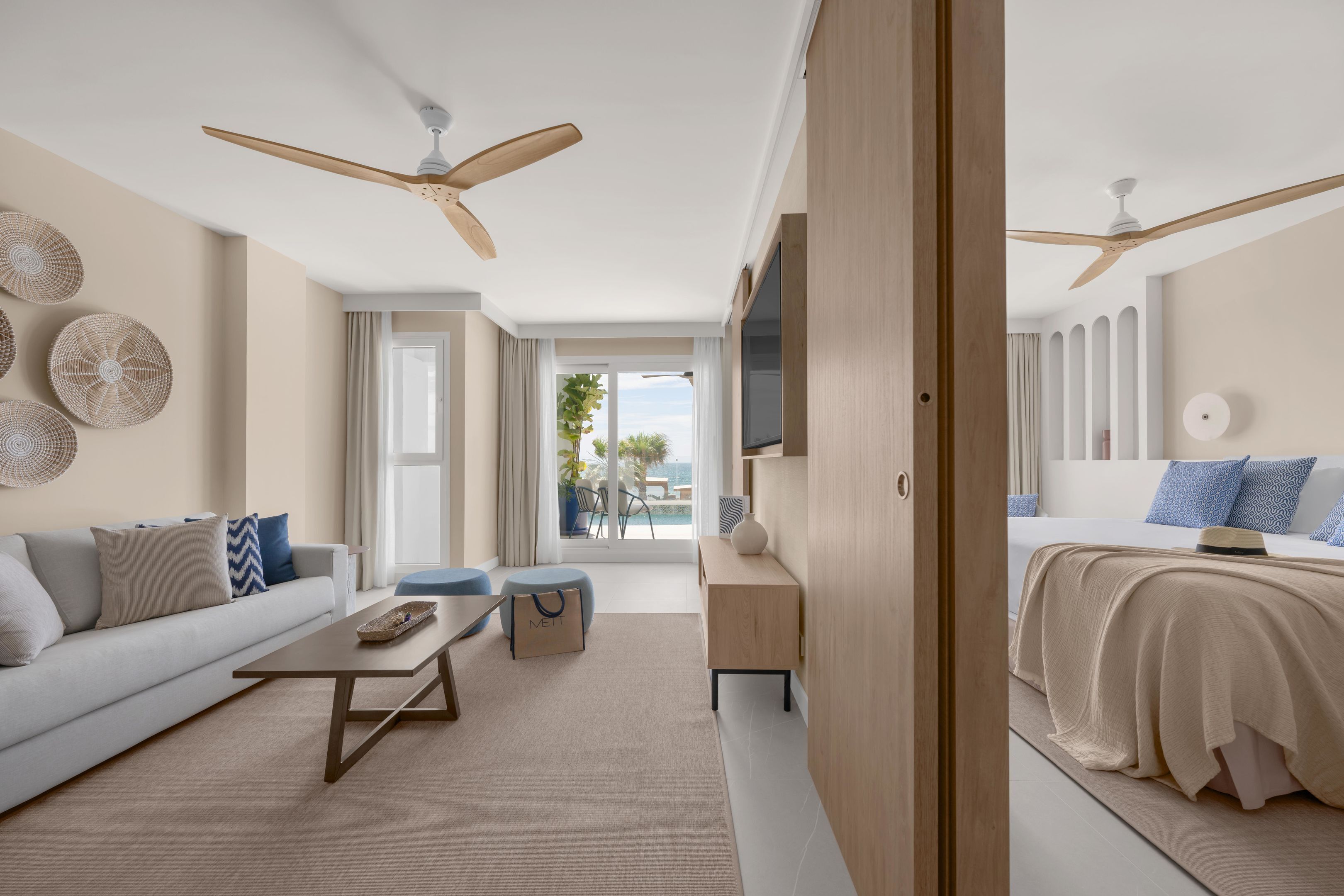 A modern hotel room with a sofa, a coffee table, a side table, a bed with a blue headboard, and a balcony with a view.