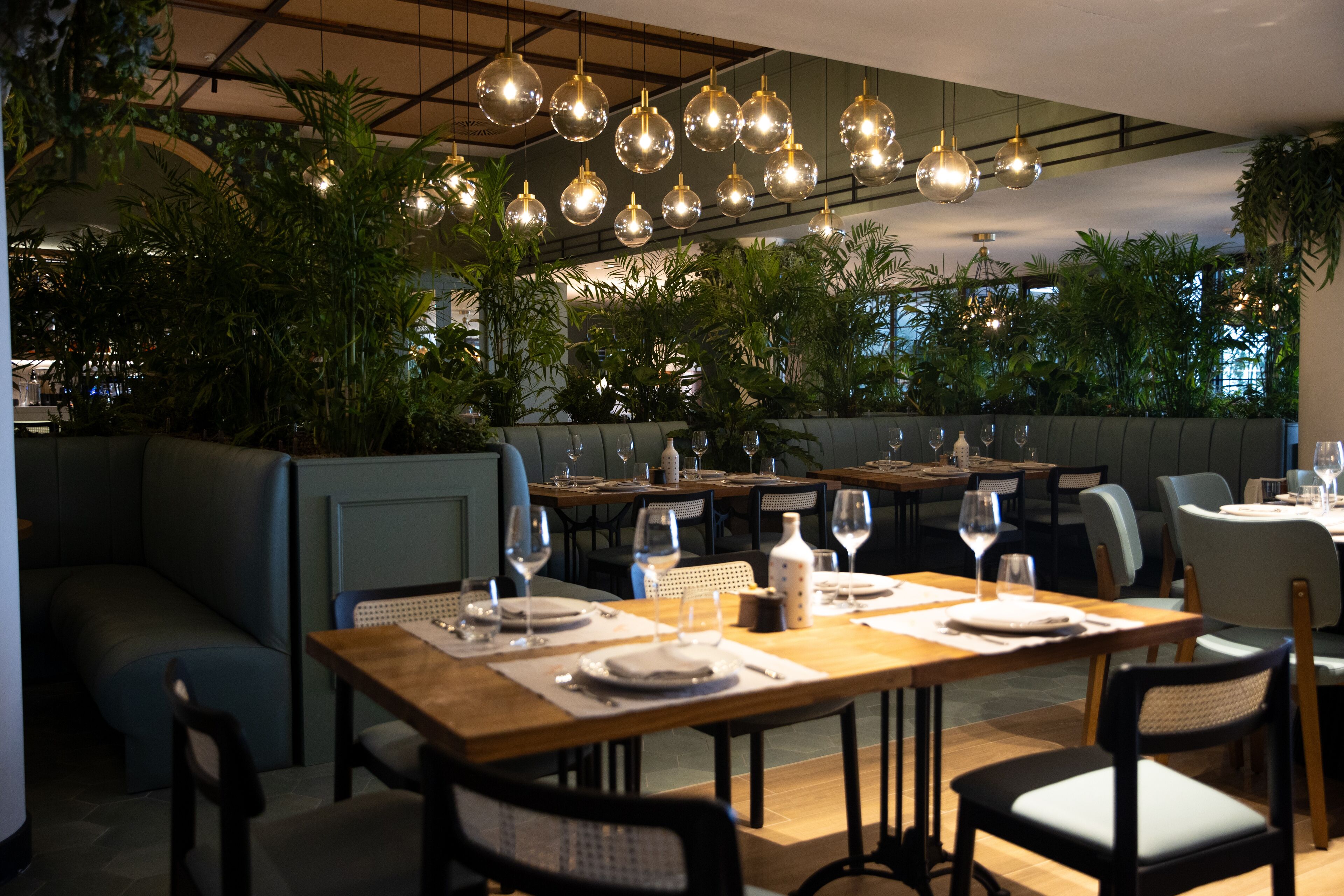 Een restaurant met meerdere eettafels gedekt voor gasten, omgeven door weelderige groene planten en moderne hanglampen.