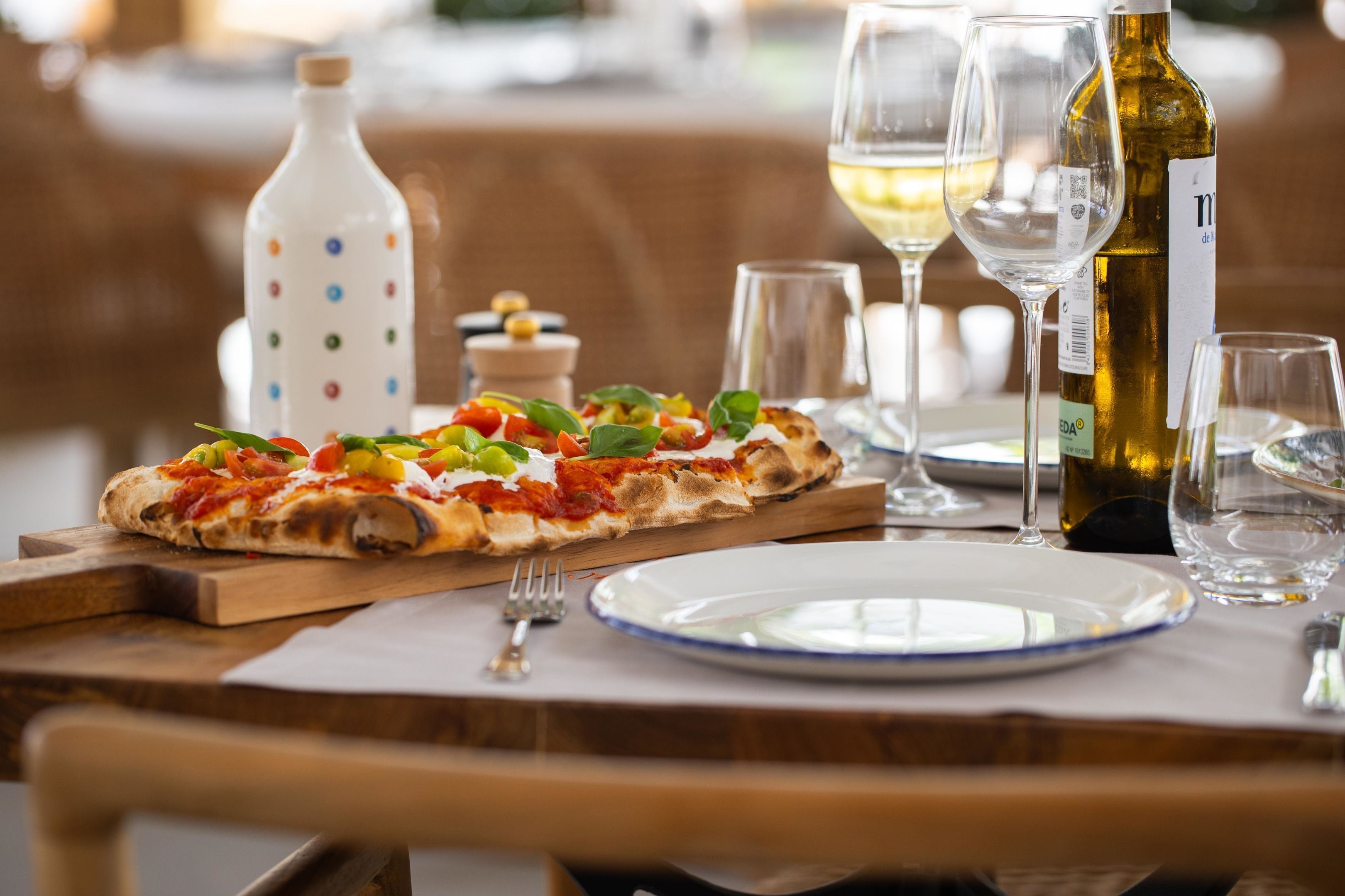 Pizza på en træplade med en gaffel og en tallerken, samt en vinflaske og glas på et bord.