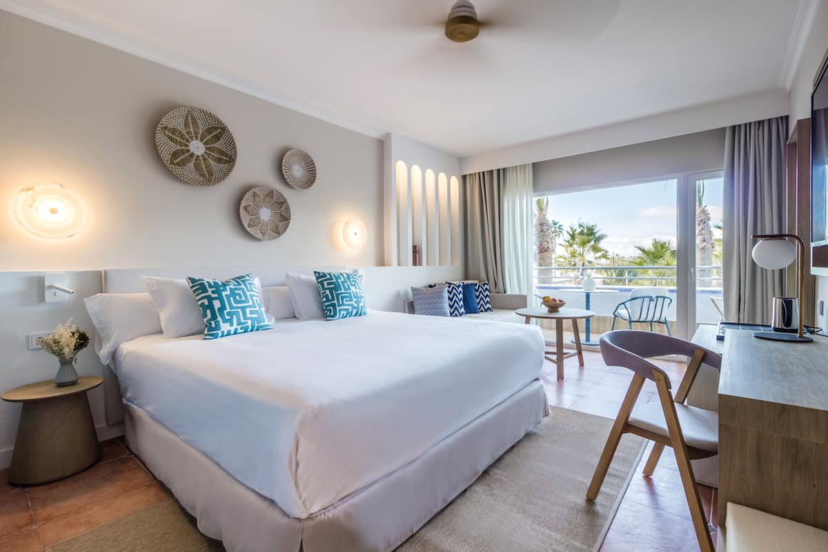 A hotel room with a large bed, a small table, a chair, and a balcony with a view of palm trees.