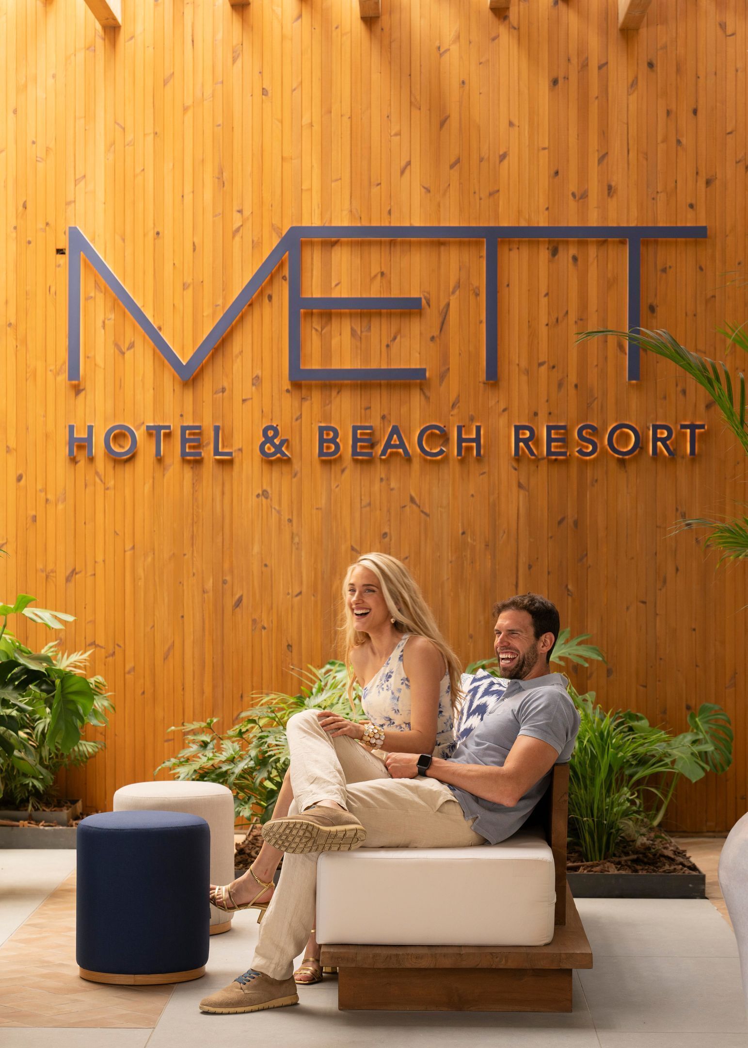 Una pareja sentada en un banco frente a un muro de madera con el letrero "METT HOTEL & BEACH RESORT".