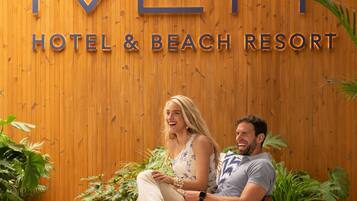 Una pareja sentada en un banco frente a un muro de madera con el letrero "METT HOTEL & BEACH RESORT".