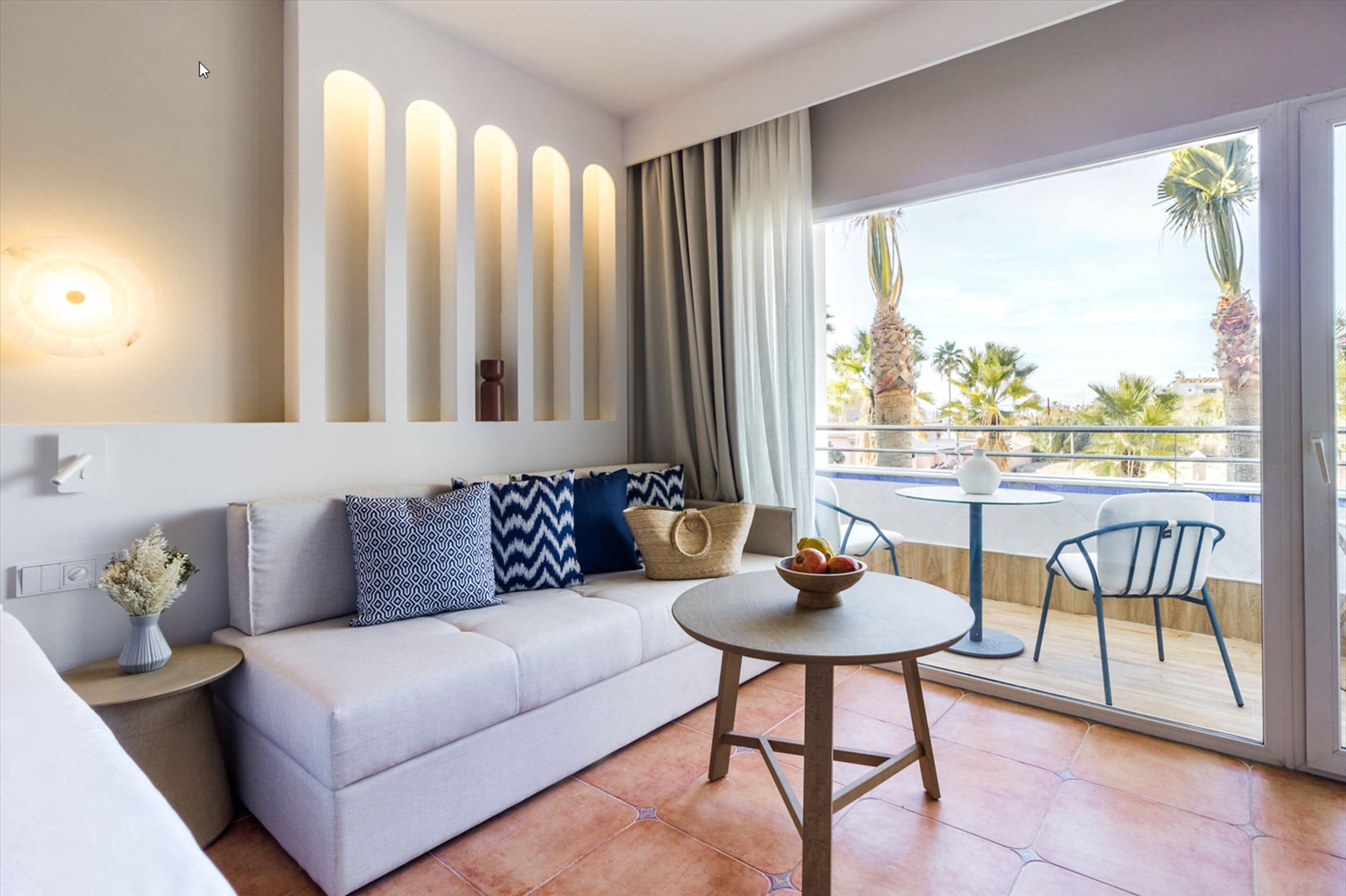 Una habitación de hotel moderna con sofá, una mesa redonda y un balcón con vistas a palmeras.