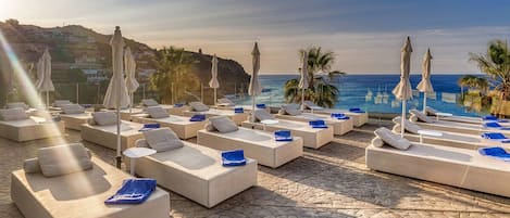 A beachside lounge area with sun loungers and umbrellas, overlooking the sea and hills.