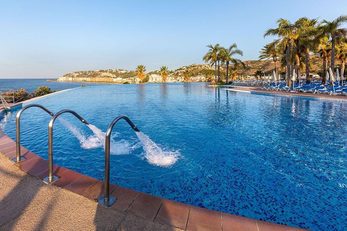 Una piscina con agua azul cristalina, rodeada de sillas de descanso y palmeras, con vistas al mar y colinas al fondo.