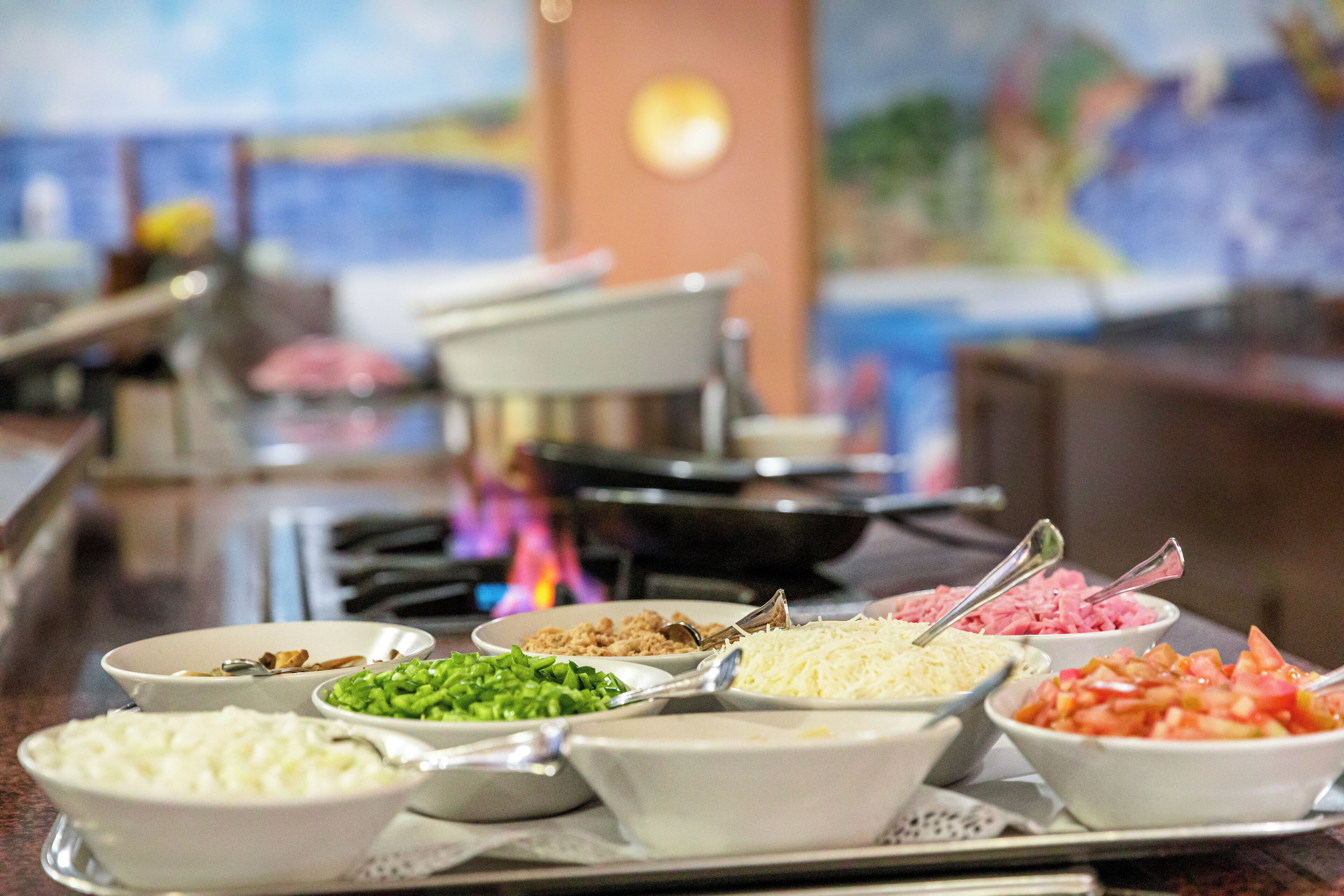 A buffet with various food items in bowls, including rice, green vegetables, cheese, and diced tomatoes.