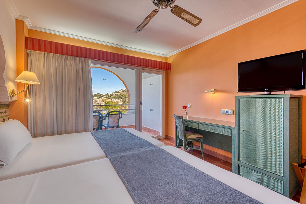 A hotel room with a bed, a desk, a television, and a view of a cityscape through a large window.