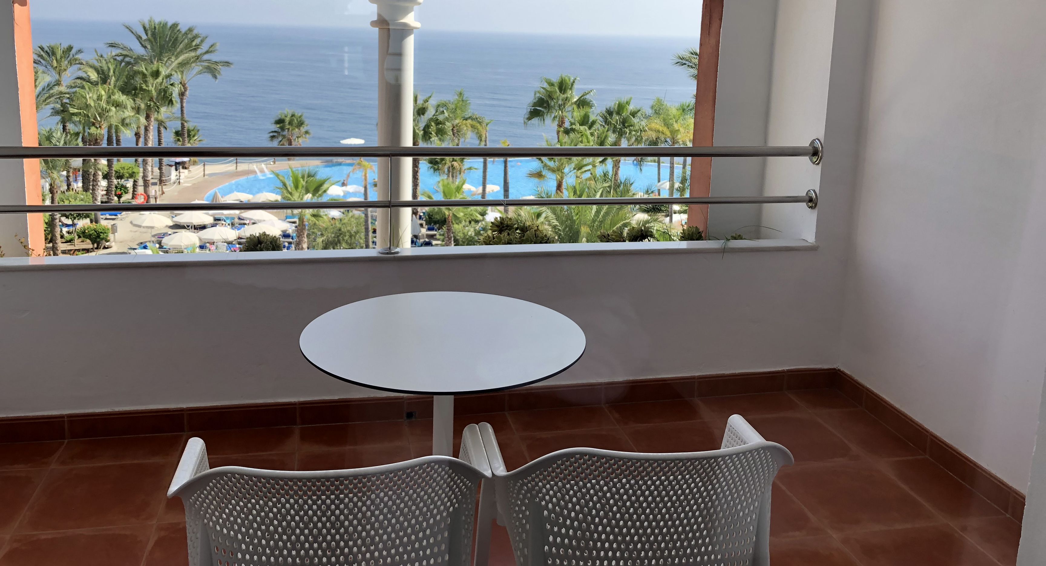 Een balkon met een witte tafel en twee stoelen, met uitzicht op een zwembad en palmbomen.