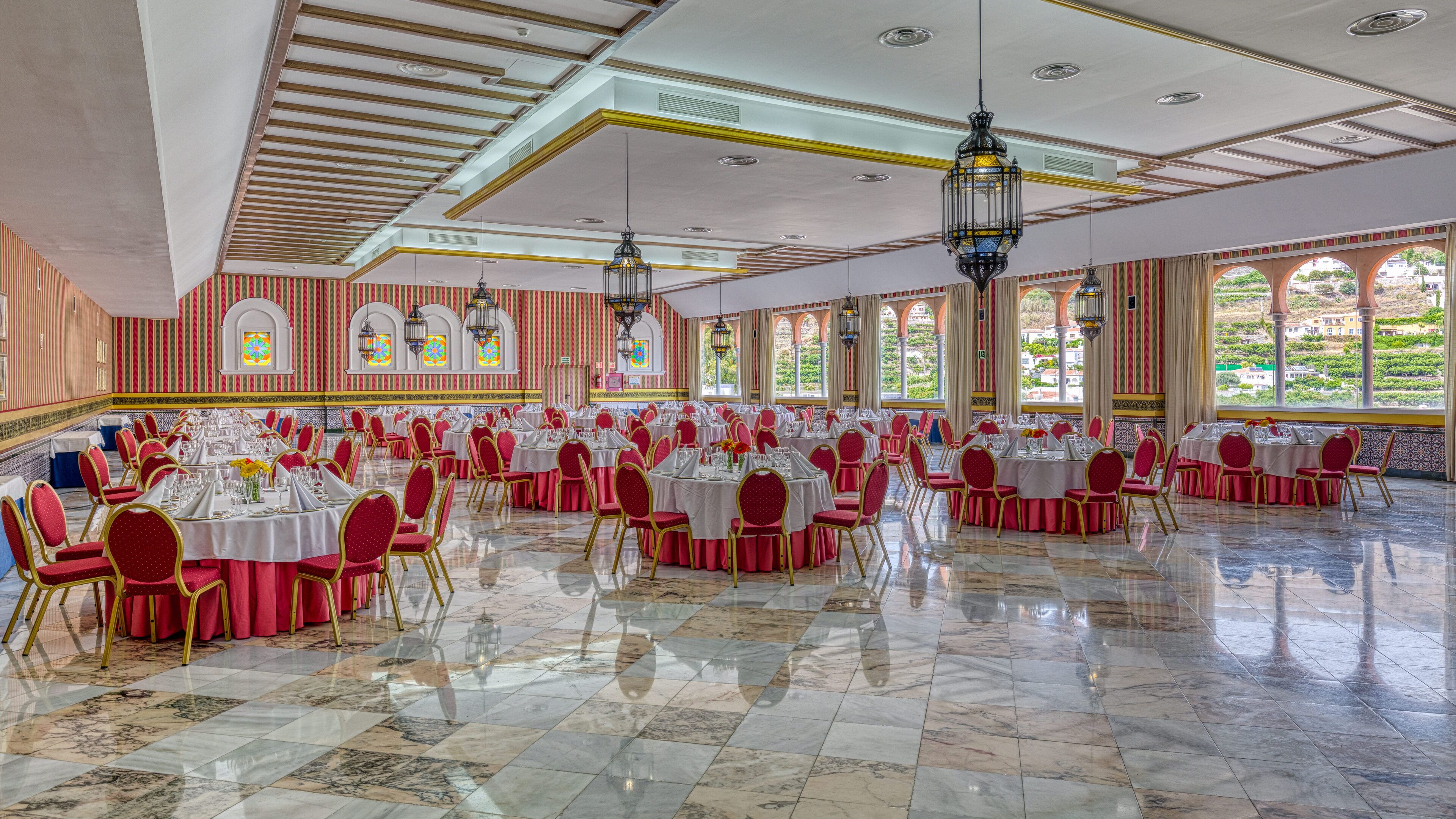 A large banquet hall with multiple round tables and chairs, high ceilings with decorative lighting, and large windows offering a view of greenery outside.