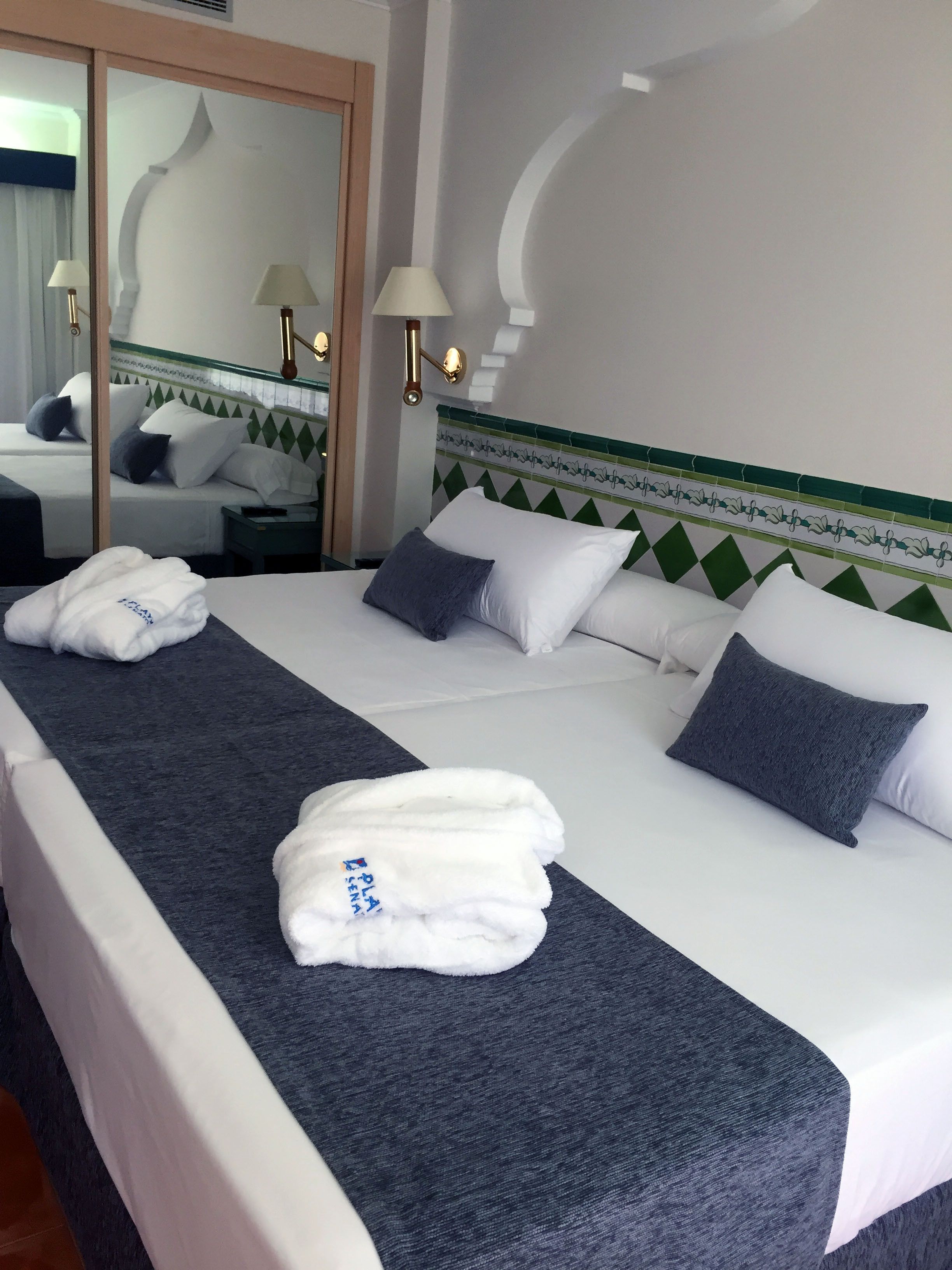 Ein Hotelzimmer mit einem ordentlich bezogenem Bett, zwei Kissen, einem gefalteten Handtuch und einem Nachttisch mit Lampe.