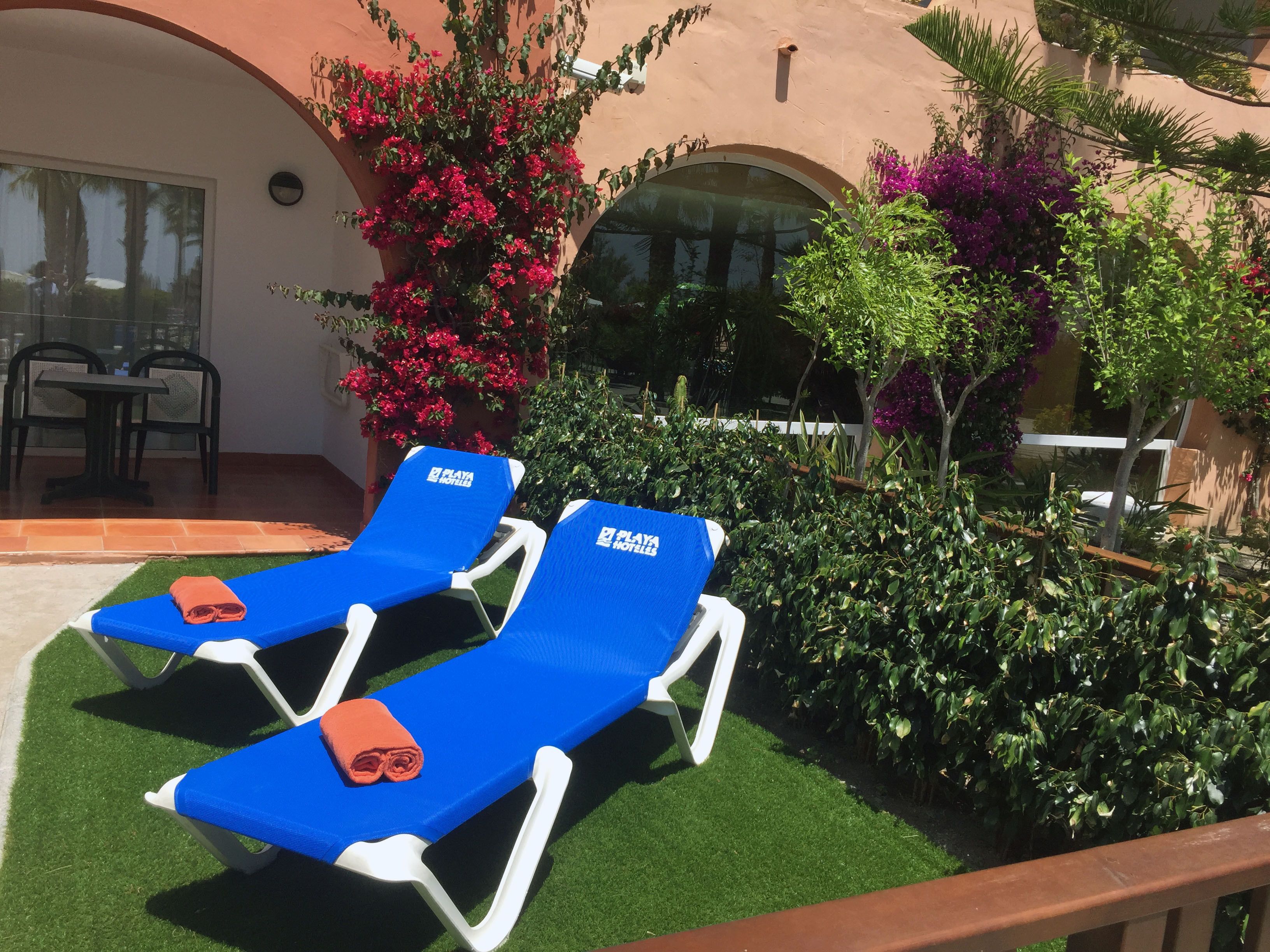 Two blue lounge chairs with towels, a patio area with artificial grass, and a variety of plants and flowers.