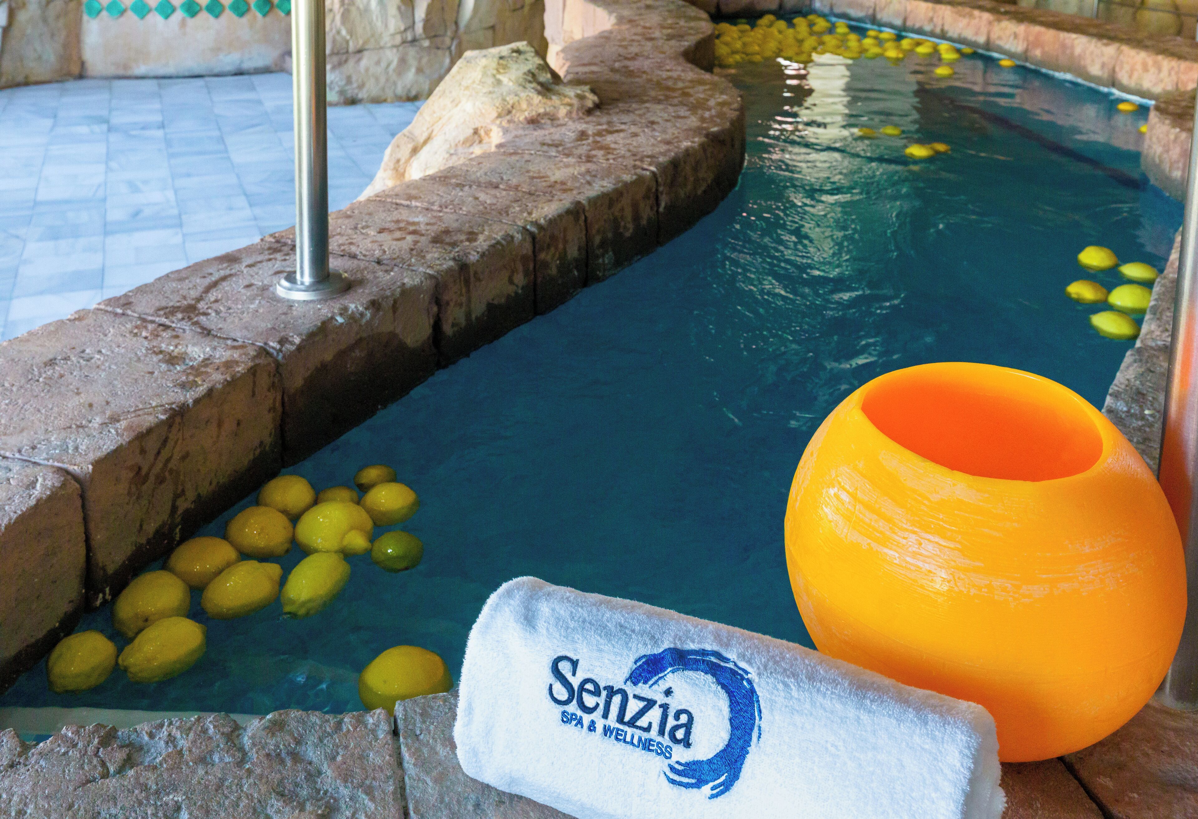 A pool with yellow floating objects, a stone edge, and a towel with 'Senzia SPA & WELLNESS' branding.