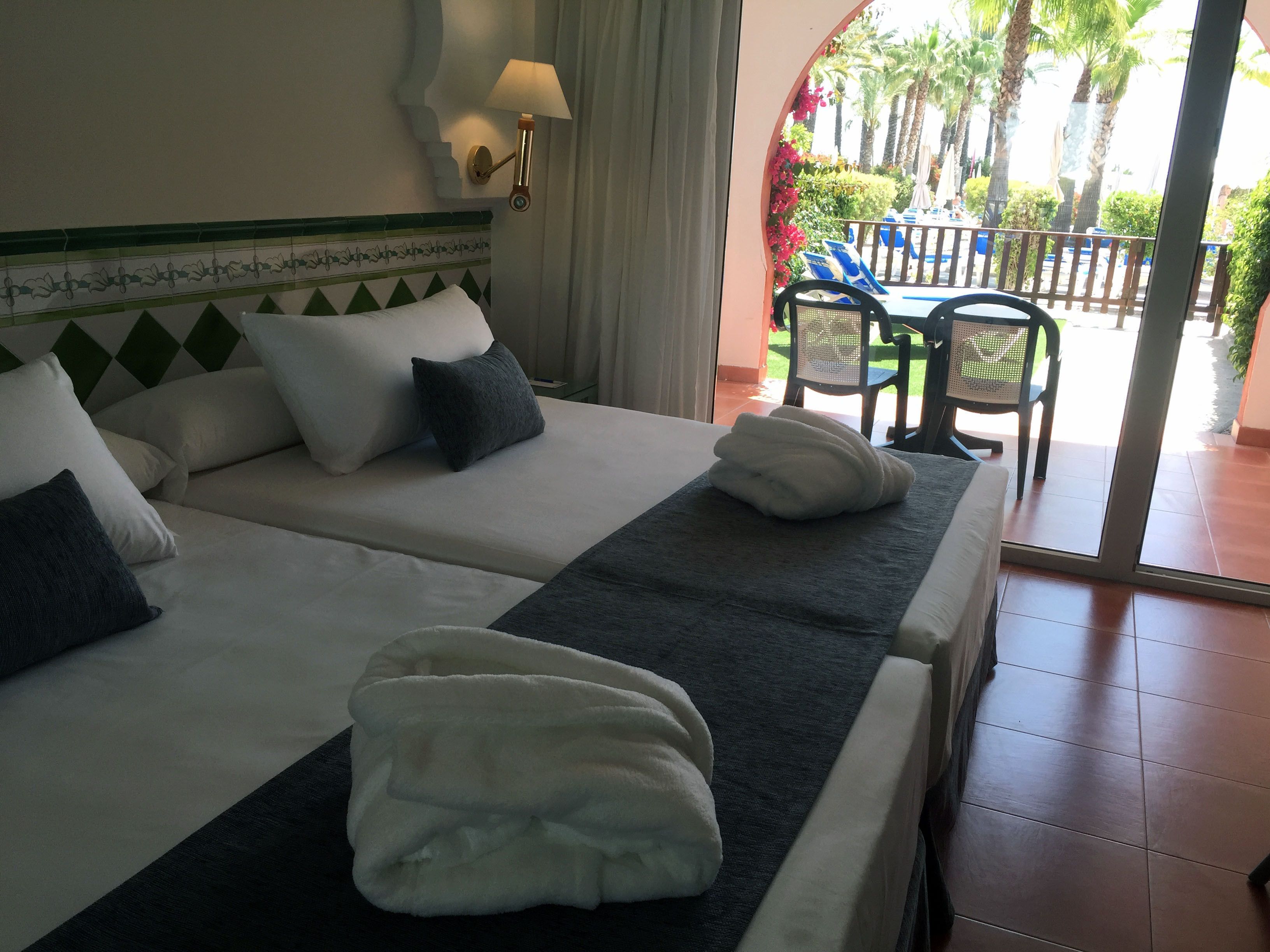 Quarto de hotel com duas camas, varanda com cadeiras e vista para palmeiras.