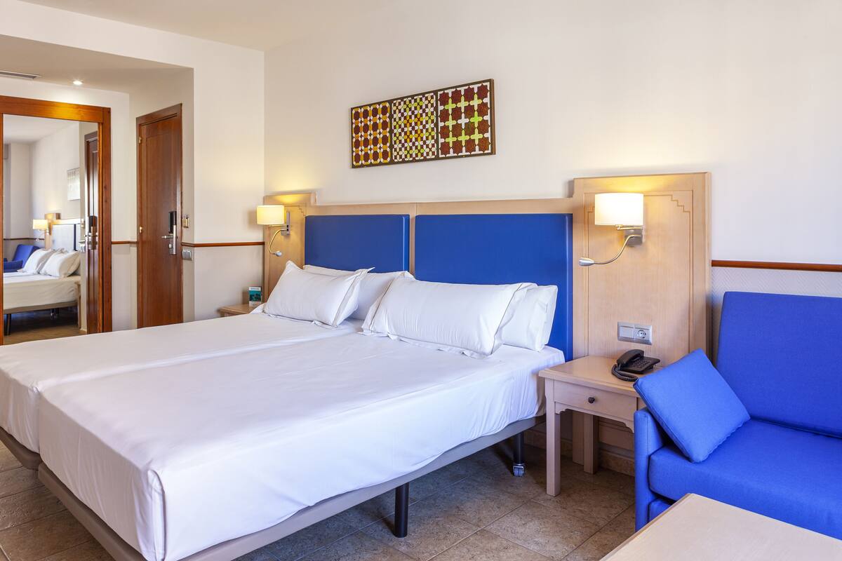 Une chambre d’hôtel avec un grand lit, un bureau avec un téléphone, une chaise bleue et un luminaire fixé au mur.