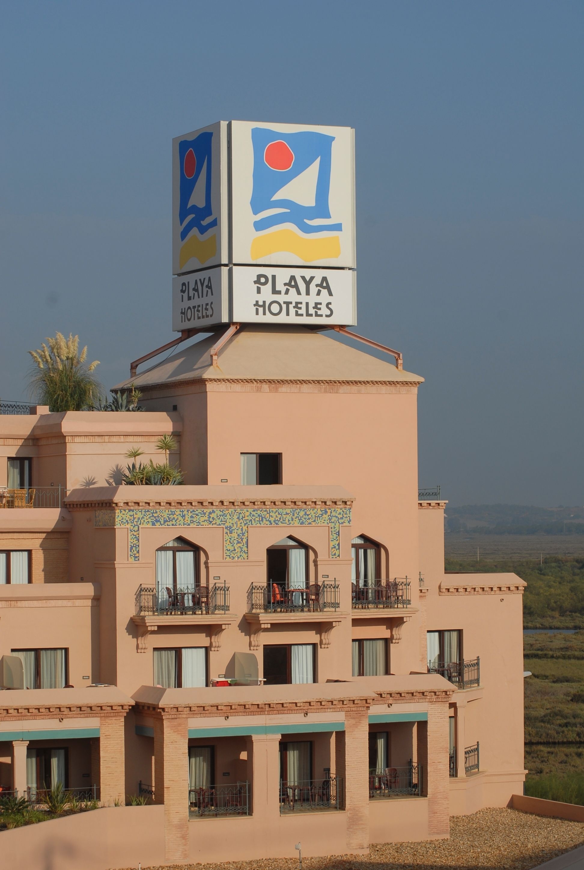 A hotel with a sign reading "PLAYA HOTELES" and a logo with a red circle and blue waves. The building has multiple balconies and a tiled facade.