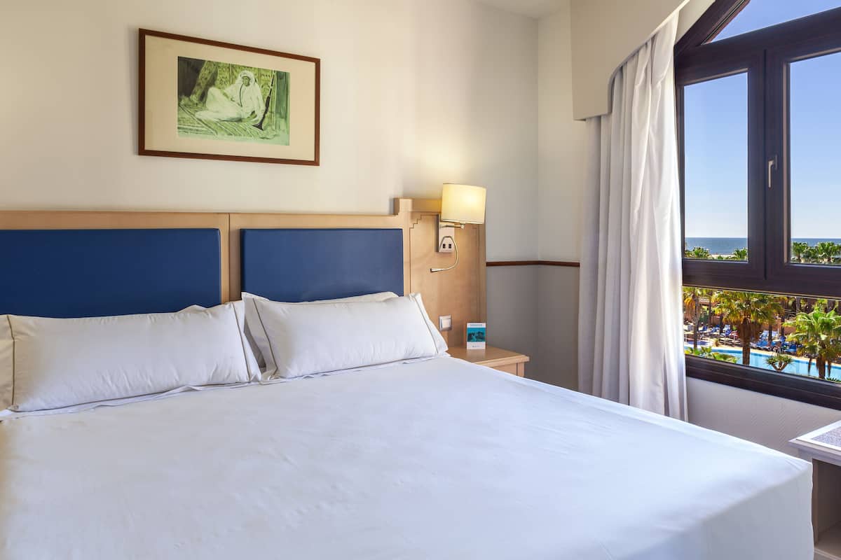 Une chambre d’hôtel avec un grand lit, une table de chevet avec une lampe, et une vue sur une piscine et des palmiers.