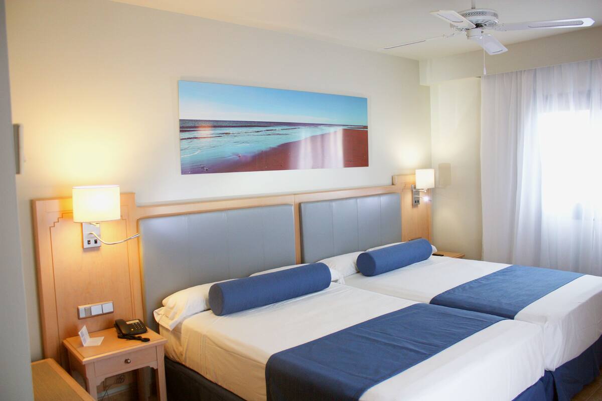 Une chambre d’hôtel avec deux lits, un tableau représentant une plage, un ventilateur de plafond et une table de chevet avec un téléphone et une lampe.