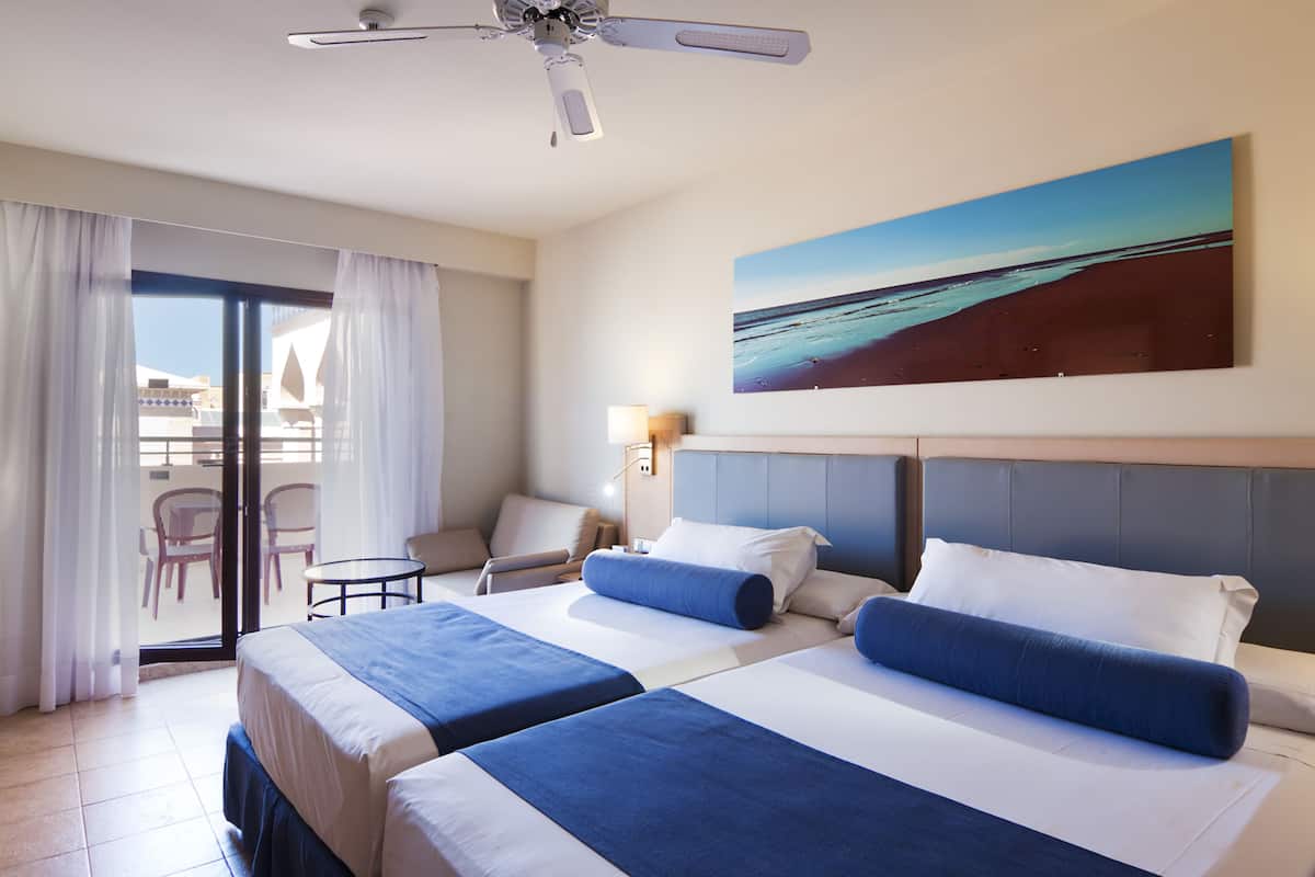 Une chambre d’hôtel avec deux lits, un ventilateur de plafond, une grande fenêtre avec des rideaux, un tableau représentant une plage et un balcon avec des chaises.